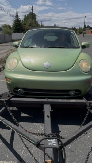 Light green VW Beetle (nicknamed Paul McCARtney) sitting awkwardly on a tow dolly in a parking lot under a partly cloudy sky. The front of the car is framed by chains and tow bars. Ironically depecting a "Living the Dream GIF"